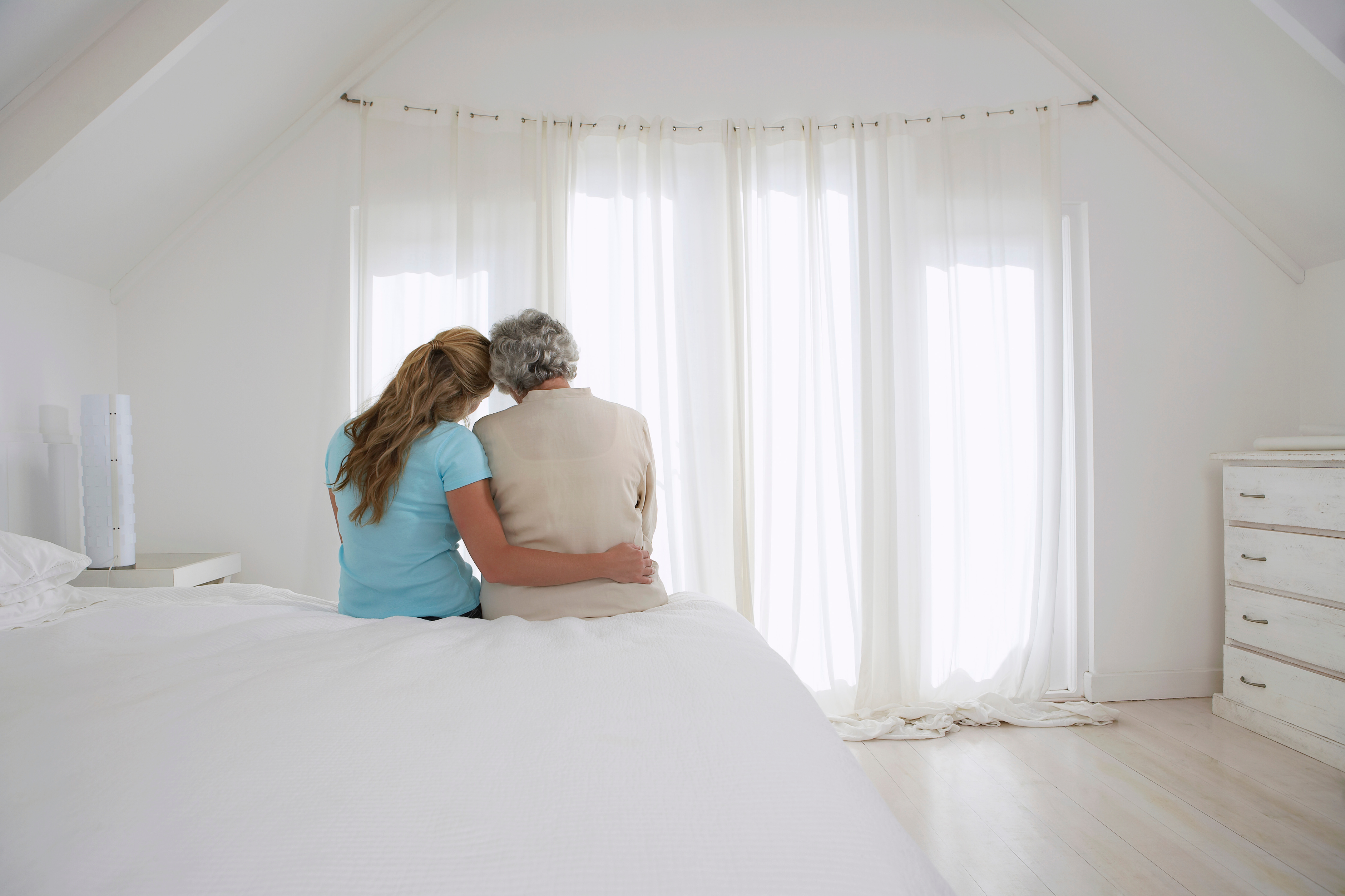 Rear view of adult daughter with her arm around mother in white bedroom